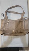 Front side of beige handbag showing gold buckle detail and two shoulder straps.