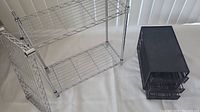 Small silver metal wire 3-shelf rack missing clips and black mesh 3-tier organizer side by side on white surface.