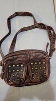 Front view of brown synthetic leather shoulder bag showing two front zip pockets with brass-tone stud decorations.