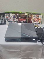 Xbox One gray console model 1540 front view with power on and disc drive visible, showing minor surface scratches. Five Xbox 360 game cases stood behind the console. Loose disc visible to the left.