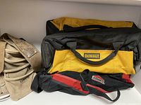 Three tool pouches/bags including a tan Kuny's roofer's belt pouch, a black and yellow DeWALT bag, and a black and red JOBMATE pouch arranged on a shelf.