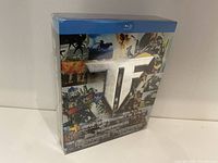 Front angled view of the sealed Transformers three movie Blu-ray set in original shrink wrap, showing silver 'TF' logo and collage of movie scenes.