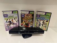 Xbox 360 Kinect sensor front view with three Kinect game cases behind it: Kinect Sports, Kinect Adventures, Kinect Sports Season Two.