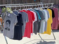 Lot of 15 graphic T-shirts hanging on a drying rack outdoors, showing various colors and prints including a gray T-shirt with a woman's portrait graphic and multiple other shirts in different shades.