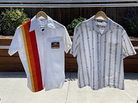 Two vintage shirts hanging on a rack outdoors, one white with bright stripes and a patch, the other white with black geometric patterns.
