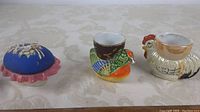 Three items photographed on a beige patterned tablecloth: bird shaped egg cups and a covered pin cushion.