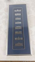 Full view of the framed pewter ornament set with seven detailed building representations arranged vertically on a blue background.
