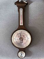 Full view of the Taylor weather station showing the three instruments: thermometer at the top, barometer in the center, and hygrometer at the bottom on a wooden frame.