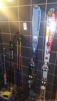 Two pairs of skis and ski poles leaning against tiled wall, two tub-shaped ski racks or bags on floor