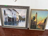 Photo showing two framed art prints on the floor, the larger one depicting a rainy street scene, the smaller showing a canal with buildings and a clock tower.