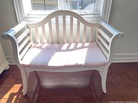 Front view of white wooden bench with pink striped cushion placed on hardwood floor near window with sunlight.