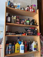 Four wooden shelves filled with various automotive fluids, sprays, and battery charger. Items include engine oils, fuel additives, and several aerosol cans.