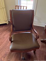 Front view of wood desk chair showing brown faux leather seat and backrest with nailhead trim, carved wood arms, and base.