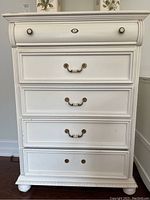 Front view of white wood dresser showing five drawers with brass handles, top drawer missing one knob and minor surface scratches.