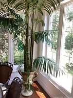 Tall faux bamboo plant with segmented stems and broad, green leaves standing inside a large decorative metal planter, near floor-to-ceiling windows.