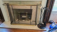Wide view showing brass fireplace screen in front of fireplace, cast iron tool stand with four tools on the right, and andirons and grate inside fireplace.