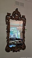 Full front view of the ornately framed mirror mounted on a wall reflecting a chandelier and window.