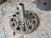 Top-down view of round metal patio umbrella base on stone floor, showing ornamental openwork.