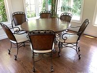 Wide angle photo showing the round wood table top with leather inlay surrounded by six metal chairs with padded white seats and leather backs, all on hardwood floor near large windows.