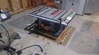 Image of Ryobi 10 inch table saw on wooden pallet in basement, showing full unit with power cord and table surface.