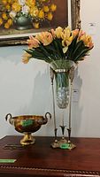 Brass pedestal bowl with swan handles and ornate brass vase with glass holder filled with faux tulip flowers displayed on wood furniture.