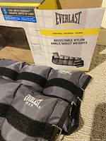 Adjustable ankle/wrist weights in grey synthetic nylon with Everlast F.I.T. logos, shown alongside the slightly torn box with product branding and details.