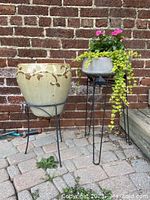 Two flower pots displayed side by side, one ceramic with floral detail and one concrete, both on metal stands, outdoor setting with brick wall backdrop.