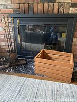 Full view of fireplace with glass doors installed, fireplace tool set with brass handles on left, and wooden slatted basket on right on the hearth.