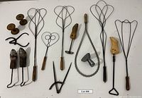 Top view showing various wire rug beaters with wooden handles, two wooden shoe stretchers with metal components, a metal iron, wooden spools, and a metal wing divider with calipers laid on a white surface.