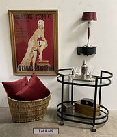 Wide shot of lot showing bar cart with items, framed 'Theatre Comic' artwork, basket with pillows, wall shelf and lamp on white wall.
