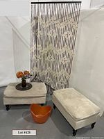 Two cream-colored matching ottomans with tufted tops and dark wooden legs shown front and side angles. Wood beaded curtain behind with geometric pattern. Orange acrylic bucket placed in front and small vase with flowers on ottoman.