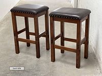 Two brown wood bar stools side by side showing front and side profiles on concrete floor with white wall background.