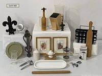Overview photo showing all items in the lot arranged on a white platform, including framed art, coffee press, grinder box, wooden utensil block, baskets, rolling pin, and various small kitchen tools.
