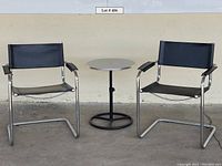 Two chrome framed directors chairs with black leather seats and backs, positioned on either side of a small round pedestal table with silver top and black base.