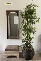 Full view of footstool, faux tree in wicker basket, and wall mirror on white wall.