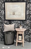 View of the large framed floral artwork, feather pillow on black metal hamper, blush pink flower-shaped side table with candle and small framed artwork on lower shelf, against a charcoal floral fabric backdrop