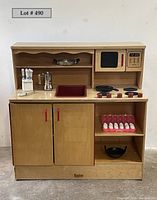 Full front view of the vintage Kaplan wooden kids play kitchen showing stove, sink, microwave, shelves, storage cabinets, and included accessories arranged on and inside the kitchen.