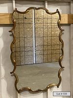 Front view of the large scalloped edge mirror with gold-tone metal frame hanging against a wooden backing structure in the warehouse.