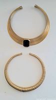 Full view of two vintage gold-tone statement necklaces displayed flat. One necklace has a wide ribbed collar with large black rectangular stone. The other is a rigid gold-tone coil necklace with rounded open ends.