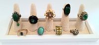 Front view of 9 vintage cocktail rings on display stand showing various green and yellow stones with gold and silver tones.