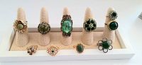 Lot of nine vintage cocktail rings displayed on a white stand. Various green stones and gold and silver settings.