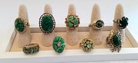 Full front view of all nine vintage green cocktail rings on beige ring holders. Shows a variety of carved, faceted, and cabochon stones with gold and silver-tone settings.