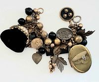 Charm bracelet laid flat showing entire length with clustered black and gold beads and charms including small black velvet mini purse with gold frame, heart charm, leaf charms, bird charm, button charm, coin charm, and a gold-tone compact charm with clear stone.