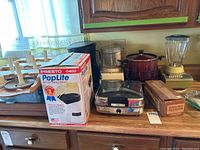 Photo showing multiple kitchen appliances arranged on a countertop including a boxed Presto PopLite hot air popcorn popper, AMC waffle iron, red crock pot, blender, and food chopper.