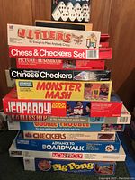 Stack of board games showing titles such as Jitters, Chess & Checkers Set, Picture Rummikub, Steel Board Chinese Checkers, Monster Mash, Jeopardy Junior Edition, Battleship, Double Trouble, Checkers, Advance to Boardwalk, Monopoly, Pig Pong