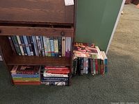 Books arranged on a wooden shelf and stacked on the floor including schoolbooks and paperbacks