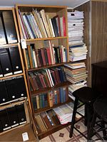 Bookshelves filled with various books including some large format music books and hymnals, with piles of paper booklets and manuals on the side near stools.