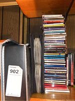 Photo of stacked music CDs showing various titles and genres, with a black cassette case and DVD boxes nearby.