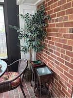 Full view showing faux tree in wicker pot next to two wicker plant stands with glass tops against a brick wall indoors.