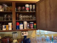 Shelf with assorted glass steins, drinking glasses, and colorful plastic cups in a wooden cabinet.